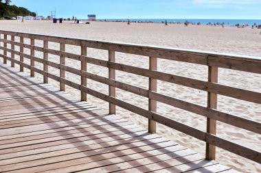 Tahta kaldırım, huzurlu kumsalın yanında yürürken tahtalarında gölgeler bırakıyor. Uzakta, renkli şemsiyeler benekli kıyılar, sakin deniz kenarı kaçışına davetiye çıkarıyor.