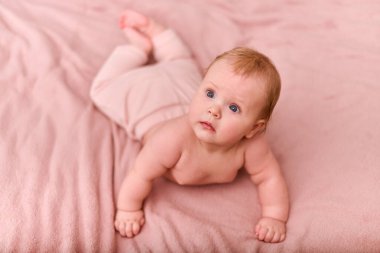 Genç, kızıl saçlı, pembe battaniyeli bir kız çocuğu dik dik bakıyor. Açık tenli, tuhaf bir ifade. Sıcak ışıklandırma, dokulu arkaplan konforu ekler