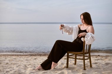 Genç bayan ressam sahilde zarif bir şekilde oturur, elinde boya fırçası tutarak. Sakin deniz ve yumuşak kum huzurlu ve ilham verici atmosfer yaratır. Sanatsal yansıma için mükemmeldir.