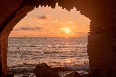 Antik tuğla kemer tarafından çerçevelenmiş huzurlu günbatımı sahnesi, Baltiysk 'teki Zapadny Kalesi' nin harabelerinin ılık renkleri ve sakin deniz atmosferini yakalıyor.