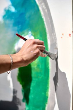 Genç, açık tenli, erkek sanatçı tuvale renkli ve soyut bir eser çiziyor. Mavi ve yeşil, yumuşak ışıklandırma ve odaklanmış perspektifle baskın sahne