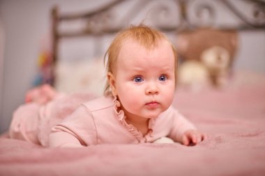 Açık renk saçlı, mavi gözlü pembe yataklı bebek. Yumuşak ışıklandırma ve yatak odası ayarları sıcak ve huzurlu bir atmosfer yaratır.