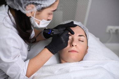 Güzellik uzmanı kuafördeki genç kadın kaşlarına kalıcı makyaj uygular. Odaklanma hassasiyet ve sanatsallık üzerinedir, sakin ortam deneyimi artırır.