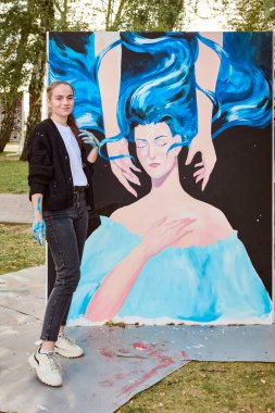 Young adult female with light brown hair, caucasian, stands beside large painting featuring blue and black tones, outdoors in park setting. Sunset light casts warm glow clipart