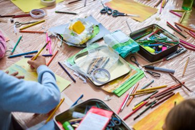 Çocuklar resim malzemeleriyle dolu bir masada resim yaparlar. Parlak renkler, yoğun ortam ve yukarıdan aşağı perspektif enerjik bir atmosfer yaratır