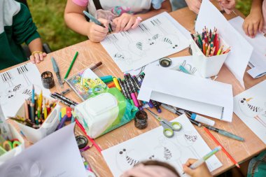 Genç eller, açık hava ortamında resim malzemeleriyle dolu masada resim çiziyor. Yeşil çimenler görünür, canlı atmosfer yaratıcılığı ve odaklanmayı arttırır.