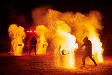 Gece gökyüzünde parıldayan iki itfaiyeci silueti, bir yangın gösterisi sırasında enerji ve ateş manipülasyonu gösteriyorlar.