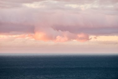 Gün batımında sersemletici bir okyanus manzarası. Canlı turuncu ve pembe bulutlar durgun mavi su ve dramatik gökyüzüne karşı.