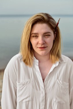 Genç bir kadın sakin bir plajda duruyor, saçlarında boya fırçası, yaratıcılık ve sükuneti çağrıştırıyor. Onun beyaz gömleği sakin denizle çelişiyor, yumuşak gün ışığı altında derin derin bir zarafet yakalıyor.