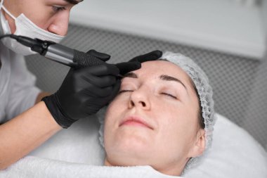 Genç bir kadın huzurlu bir kaplıcada kaş makyajı yaptırıyor. Güzellik teknisyeni, eldiven takıyor, dikkatle odaklanıyor, sakin, profesyonel atmosfer yaratıyor.