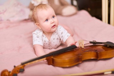 Sarı saçlı küçük kız pembe yatağa uzanmış, garip bir şekilde kemana uzanıyor. Konforlu yatak odası ayarları, sıcak ışıklandırma huzurlu ve neşeli atmosferi geliştirir.