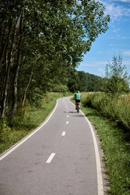 Yaşlı yetişkin, mavi gökyüzünün altında yemyeşil yemyeşil ile çevrili sakin park yolu boyunca bisiklet sürüyor.