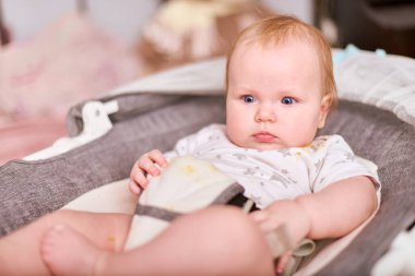 Yüksek sandalyede açık renk saçlı, yıldız desenli tulum giyen bir bebek. Yumuşak ışıklandırma, rahat ortamda nötr tonlar. Çocuk rahat, meraklı ve hoşnut görünüyor