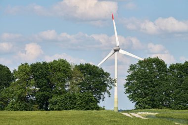 Rüzgâr türbini kırsal beton yolun sonunda duruyor. Parlak mavi gökyüzünün altındaki yemyeşil ağaçlar çerçeveli. Doğanın ve yeniliğin buluşunu simgeliyor.