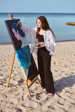 Genç kadın ressam okyanus kenarında tuval üzerinde çalışarak deniz manzarasından ilham alıyor. Odaklanmış ifadesi ve sakin arka planı yaratıcılığı ve sükuneti vurguluyor.