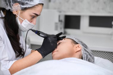 Yetenekli teknisyen, sakin salondaki genç bayana kalıcı kaş makyajı yapıyor. Siyah eldivenlerle, profesyonel aletler kullanılıyor, hassas ve rahatlatıcı ortamlarda bakım sağlanıyor.