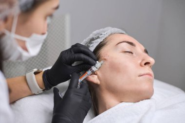 Genç bir kadın klinik çevrede profesyonel olarak yüz enjeksiyonu yaptırıyor. Prosedür estetiği, duyarlılığı, dikkati ve dingin atmosferi arttırır.