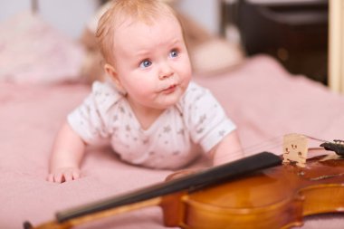 Sarı saçlı küçük kız pembe yatağa uzanmış, garip bir şekilde kemana uzanıyor. Konforlu yatak odası ayarları, sıcak ışıklandırma huzurlu ve neşeli atmosferi geliştirir.