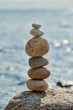 Sakin deniz kıyısında kayaların üzerinde dengelenmiş pürüzsüz taşlar yığını, denge ve sükuneti sembolize eder, barışçıl ve meditasyonel atmosferi çağrıştırır.