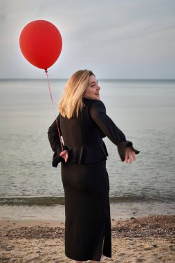 Zarif siyah elbiseli genç bir kadın, elinde kırmızı balonla sakin bir sahilde duruyor. Huzurlu deniz manzarası ve nazik gülüşü sakin ve içsel bir hava yaratıyor.