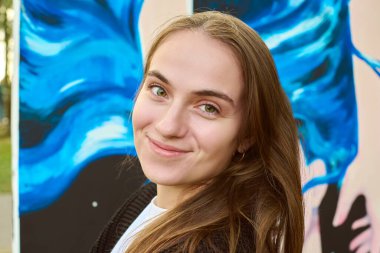 Açık kahverengi saçlı ve Kafkas kökenli genç bir kadın açık hava resminin yanında gülümsüyor. Parlak doğal aydınlatma onun neşeli ifadesini vurgular.