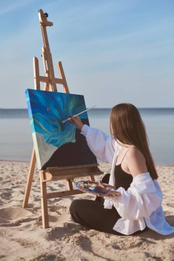 Young woman paints abstract ocean scene on canvas, seated on sandy beach. Tranquil sea setting enhances creativity, symbolizing inspiration and serenity clipart