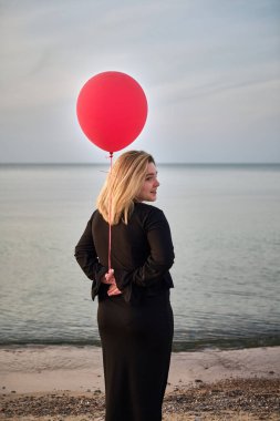 Genç kadın, alacakaranlıkta dingin bir sahil kenarında elinde canlı kırmızı bir balon tutuyor. Sakin su ve pastel gökyüzü huzuru ve ince neşeyi çağrıştırır.