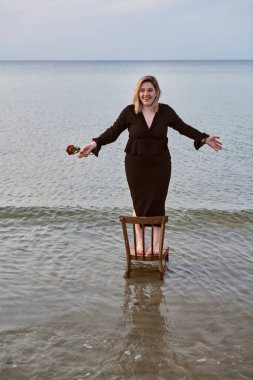 Siyah kıyafetli genç bir kadın güvenle okyanustaki sandalyede duruyor, elinde gül var. Sahne, doğanın enginliği arasında denge, cesaret ve sükunet temaları uyandırır.