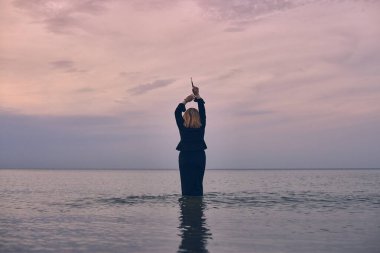 Woman stands waist-deep in tranquil ocean at sunset, raising her arms in contemplation. Peaceful setting, with pastel skies, evokes sense of freedom and introspection clipart