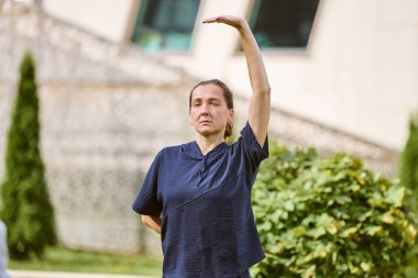 Kahverengi saçlı olgun bir dişi açık havada qigong oynar. Odaklanmış ifade, nazik hareketler. Yeşillik ve yumuşak ışıklandırma sakinliği arttırır