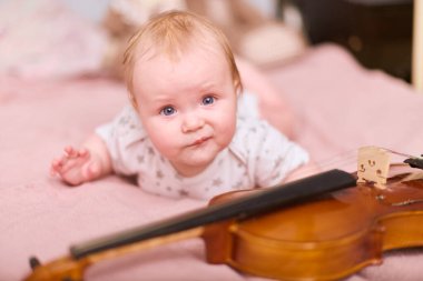 Sarı saçlı küçük kız pembe yatağa uzanmış, garip bir şekilde kemana uzanıyor. Konforlu yatak odası ayarları, sıcak ışıklandırma huzurlu ve neşeli atmosferi geliştirir.