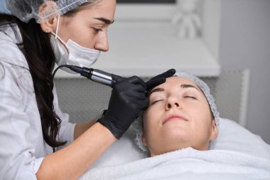 Genç bir kadın huzurlu bir kaplıcada kaş makyajı yaptırıyor. Güzellik teknisyeni, eldiven takıyor, dikkatle odaklanıyor, sakin, profesyonel atmosfer yaratıyor.