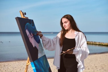 Genç kadın ressam okyanus kenarında tuval üzerinde çalışarak deniz manzarasından ilham alıyor. Odaklanmış ifadesi ve sakin arka planı yaratıcılığı ve sükuneti vurguluyor.