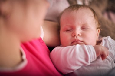 Sarışın genç bir kadın açık renk saçlı uyuyan bir bebek beşiği. Sıcak ışıklandırma, yumuşak arka plan rahatlatıcı bir atmosfer yaratır. Pembe, bej renkler sahneye hakim.