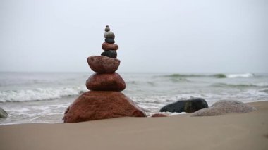 Sakin denizin yanındaki kumlu kumsalda dikkatlice dengelenmiş taş kule. Dalgaların nazikçe dalgalandığı bulutlu gökyüzü, sakin bir atmosfer yaratıyor. Doğal renk tonları huzur dolu sahneleri canlandırır
