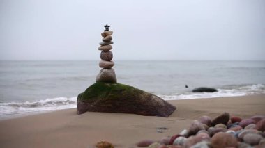 Dengeli taşlar, sisli kıyı şeridine karşı yosun kaplı kaya üzerinde dikey kule oluştururlar. Bulutlu gökyüzü ve sakin deniz sakin bir atmosfer yaratır. Yumuşak çakıl taşları kumlu ön planda dağılır