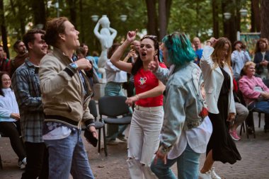 Kaliningrad, Rusya - 11.08.2024 - Farklı etnik kökenlere sahip genç yetişkinler canlı park sahnesinde enerjik bir şekilde dans ediyorlar. Hava bulutlu, arka planda heykeller ve ağaçlar var.