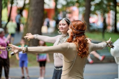 Kaliningrad, Rusya - 10.08.2024 - Açık havada kabile dansı yapan iki genç kadın. Kafkas ve zincirli kızıl saçlı dansçı. Arka planda seyirci ve ağaçlar