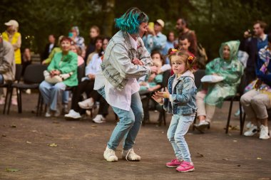 Kaliningrad, Rusya - 11.08.2024 - mavi saçlı ve çocuklu yetişkin bir kadın, ikisi de beyaz, parkta neşeyle dans ediyor. Sıradan kalabalık, doğal ışık, yeşil zemin