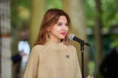 Kaliningrad, Rusya - 10.08.2024 - bej kazak giyen genç beyaz kadın parkta neşeyle şarkı söylüyor. Mikrofon önplanı, arka planda bulanık ağaçlar, doğal ışıklandırma