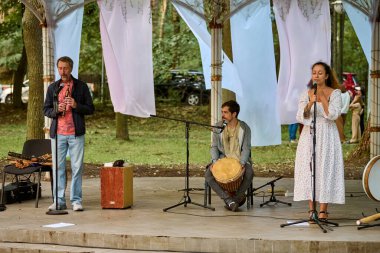 Kaliningrad, Rusya - 10.08.2024 - Ağaçların arasında üç yetişkin müzik yapıyor. Manzara açık havada, huzurlu, sıcak aydınlatma ve doğal çevreyle