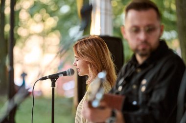 Kaliningrad, Rusya - 10.08.2024 - Genç kadın mikrofonda şarkı söylüyor, erkek gitar çalıyor. Güneş ışığı ağaçların arasından süzülür ve sıcak bir atmosfer yaratır.