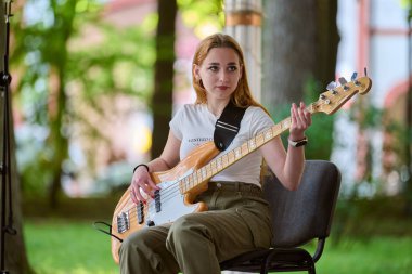 Kaliningrad, Russia - 10.08.2024 - Young female musician plays bass guitar in sunlit park, surrounded by greenery. Casual setting, focused expression clipart