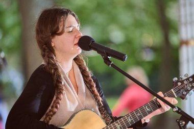 Kaliningrad, Rusya - 11.08.2024 - örgülü, şarkı söyleyen ve parkta gitar çalan beyaz bir kadın. Doğal ışıklandırma onun odaklanmış ifadesini vurgular. Arkaplanda yeşil ağaçlar