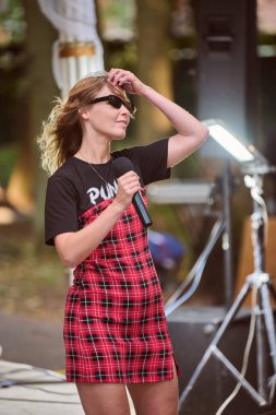 Kaliningrad, Rusya - 11.08.2024 - Kırmızı ekose elbiseli ve güneş gözlüklü genç kadın park sahnesinde tutkuyla şarkı söylüyor. Çevreleyen yeşil ağaçlar