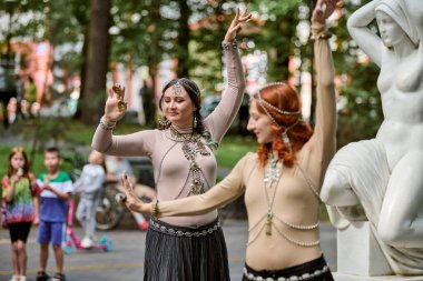 Kaliningrad, Rusya - 10.08.2024 - Açık havada kabile dansı yapan iki genç kadın. Kafkas ve zincirli kızıl saçlı dansçı. Arka planda seyirci ve ağaçlar