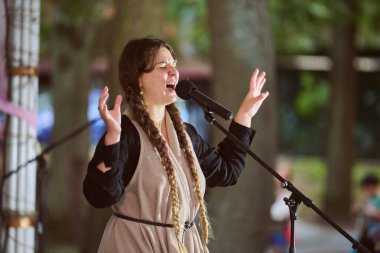 Kaliningrad, Rusya - 11.08.2024 - örgülü genç bayan şarkıcı yeşil park ortamında doğal ışık altında tutkuyla şarkı söylüyor. Ağaçlar dingin bir zemin oluşturur