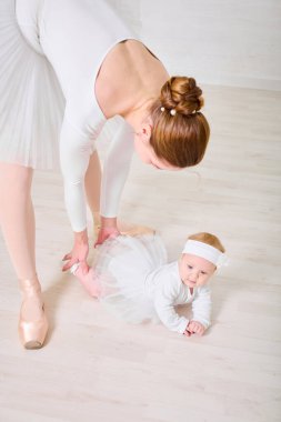 Kızıl saçlı genç bayan balerin, bembeyaz giyinmiş, açık ahşap zeminde zarif bir şekilde küçük bir kızın üzerine eğiliyor. Parlak ışık dingin, samimi bir atmosfer yaratır.