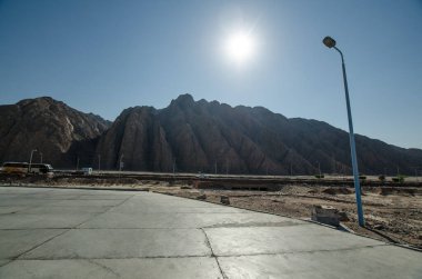 Boş tatil köyü otoparkı aydınlık, berrak gökyüzünün altında geniş bir dağ manzarası sunuyor. Güneş beton üzerine uzun gölgeler bırakır, uzak ve sakin bir çevreyi vurgular. Stark, minimalist sahne