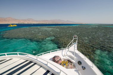 Yat yayı açık gökyüzünün altındaki canlı Kızıldeniz resiflerine bakıyor. Turkuaz sular çeşitli deniz canlılarını ortaya çıkarır. Uzak dağlar sahnelere huzur katar